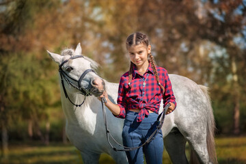 A beautiful teenage girl stands and smiles next to a white pony in autumn