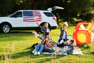 American family spending time together. With USA flags against big suv car outdoor.