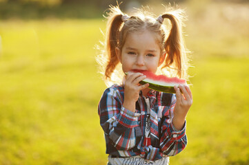 Little girl wear checkered shirt with ponytails on her head, eat watermelon.