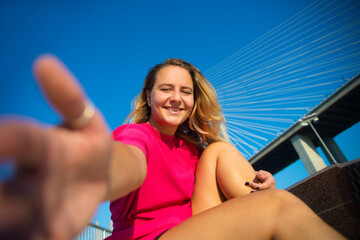 Beautiful young woman taking selfie. Pretty smiling girl in crimson dress with skateboard sitting near bridge, taking photo of herself. Smiling at camera. Sport, hobby, active lifestyle concept