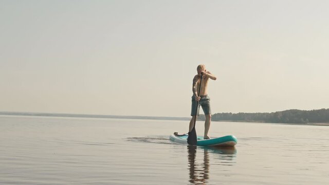 Full shot of young Caucasian man standing on sup board in lake for first time, losing balance and falling in water