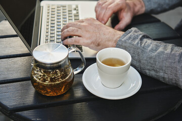 Caucasian man faceless works on a computer in a street city cafe. Herbal tea is on the table.