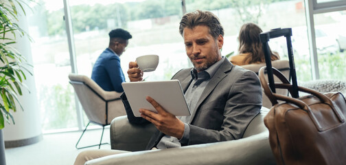 Businessman Using Digital Tablet and Drinking Coffee while Waiting for his Flight in the Airport VIP Lounge. 

Smiling business man sitting in armchair and working on tablet while holding cup of tea.