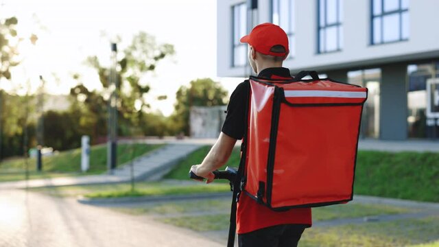 Unrecognizable young courier rides on his e-scooter. Deliver male parcel delivery service guy rides an electric scooter down an empty sidewalk with red backpack down the street at sunset