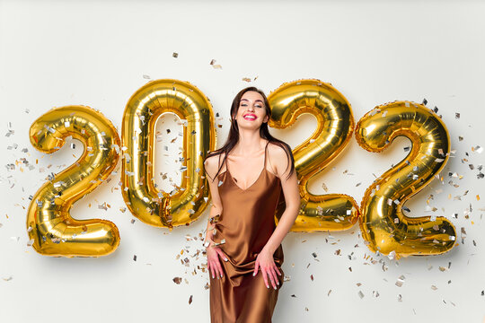 Happy young brunette woman near golden 2022 balloons on white background. New Year celebration. Wearing fashionable glamor dress. Confetti background. Smiling, Laughing - Powered by Adobe