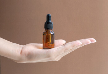 Woman's hand holds mock up of glass dropper bottle on brown background in palm of her hand. Concept of beauty industry, natural cosmetics