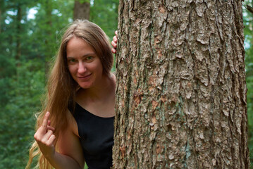 A blonde Caucasian lady with long hair looking from behind a tree trunk showing a middle finger