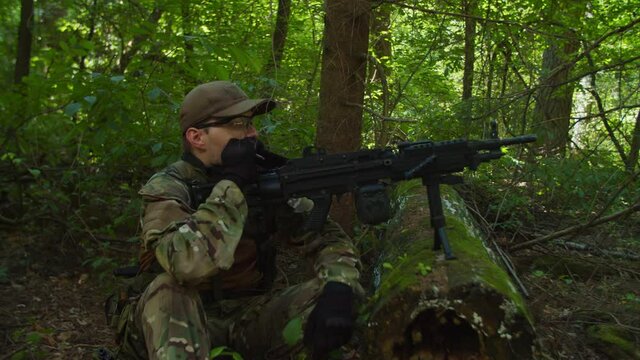 Military guard machine gunner in combat gear observing field of view in forest, smoking, resting while group of brave soldiers in camouflage clothing resting in halt, preparing rations to eat outdoor
