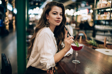 Portrait of trendy dressed diva woman looking at camera during cocktail time in local bar restaurant, attractive Caucasian customer with alcohol beverage posing during weekend leisure in public cafe