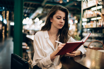 Caucasian female student with education textbook making informative notes while learning indoors, intelligent young hipster girl planning organisation during weekend studying in cafe interior