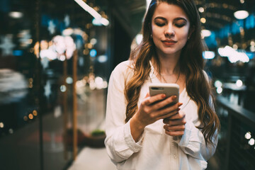 Youthful female blogger using cellular device for online networking during weekend leisure,...