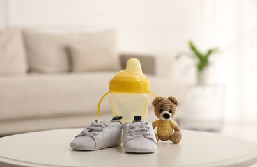 Baby bottle and shoes near toy bear on white table in room. Maternity leave concept - obrazy, fototapety, plakaty