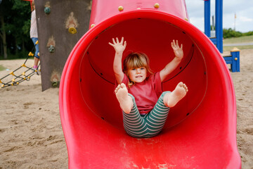 Little preschool girl playing on outdoor playground. Happy toddler child climbing and having fun with summer outdoors activity. Girl slinding down the slide. Children having fun.
