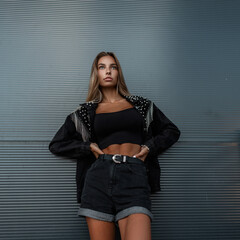 Stylish young girl model with fashion denim jacket in black jeans shorts stands near a black wall outdoors