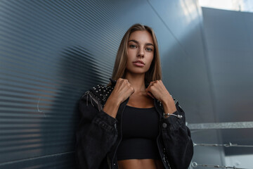 Stylish beautiful young woman with a caucasian face in fashionable black denim clothes with a top T-shirt stands near a modern metal dark building in the city