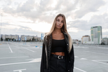 Beautiful young woman in a fashionable black denim jacket with rhinestones in stylish black jeans walks on the street in the parking lot