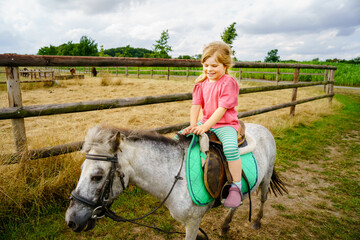 Little preschool girl riding a pony on a farm. Happy lovely child practicing horseback riding....