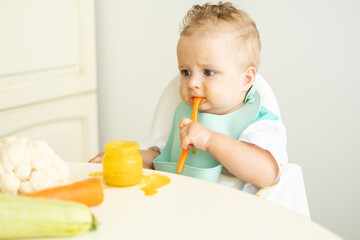 funny baby boy in bib eating vegetable puree with spoon sitting in child chair. Child learn to eat...