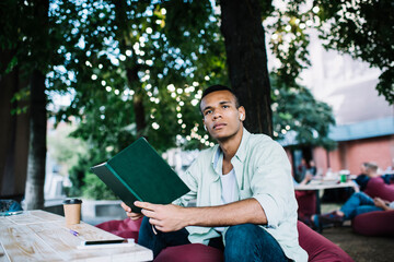 Dreamy young guy enjoying reading book while sitting in summer cafeteria
