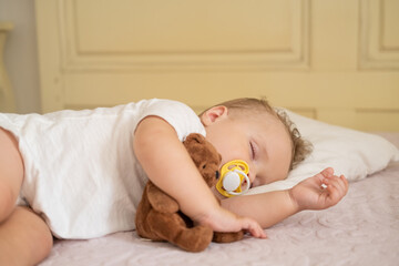 cute baby boy with nipple sleeping on bed at home. child hugging teddy bear. 