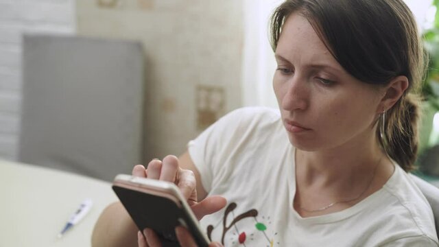 young woman measures her body temperature orally, passes an online medical consultation using a video call on a mobile phone. A sick woman with coronavirus fever, holding a thermometer