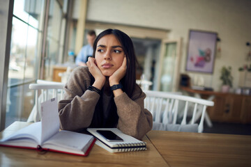 Middle Eastern female student with education notepad looking away feeling bored and tired, South ASian blogger learning and studying in cafe interior unhappy with time for organization planning