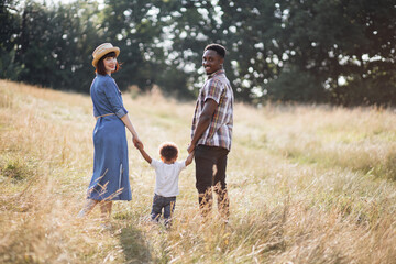 Back view of little african boy holding hands with caring parents while standing on summer nature. Caucasian woman and african man smiling and looking at camera. Family and love concept.