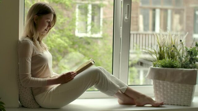 Young adult girl with book in hands reads while sitting by opened window on sill. Female reader in cozy home at reading hobby. Rest with drama story literature stress relief. Woman sits on windowsill.