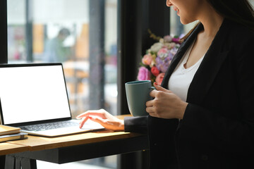 Closed up with young businesswoman holding a hot coffee cup while sitting in front of a white blank screen laptop.