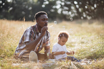 Handsome african american man blowing soap bubbles during summer picnic with cute son. Happy father spending time with his little child on fresh air.
