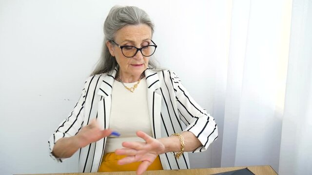 Senior grey haired businesswoman in striped jacket with eyeglasses is writing something on a paper in here office sitting at the desk. Happy retirement, employment and labor