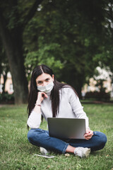 A young girl in a protective mask works in the park on a laptop and with a phone