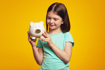 Little attractive girl holding money box in shape of pig near ear, looking happy, isolated over yellow background