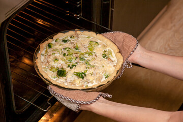 Woman takes an open shortbread pie with chicken and broccoli, sprinkled with cheese out of the oven in her kitchen