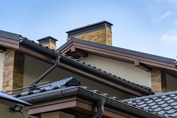 Brick house with drainage system on metal roof. Сhimney pipes is finished externally with natural stone. Roof gutter pipeline system. House rain gutter with holders and downspout pipe against blue sky