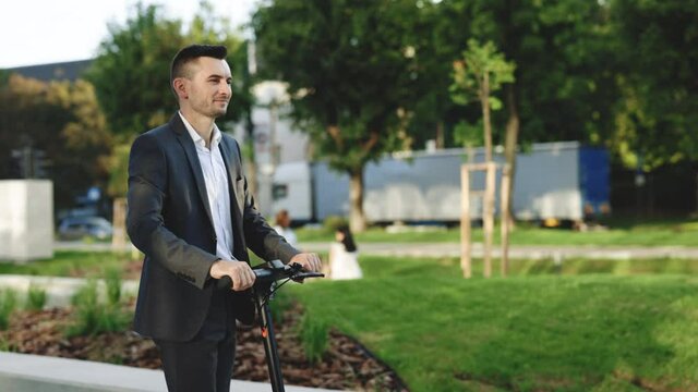Slow-motion side-view of caucasian businessman riding on electric scooter along sidewalk on warm sunny day. Modern transportation gadget and popular futuristic device among young people.