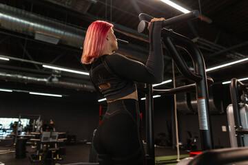 Fototapeta na wymiar Beautiful young fitness woman in black sportswear does exercises and pumps back muscles in the gym