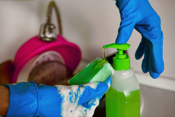 Close-up of hands in rubber gloves holding a sponge and squeezing out dishwashing liquid. Green detergent. - obrazy, fototapety, plakaty