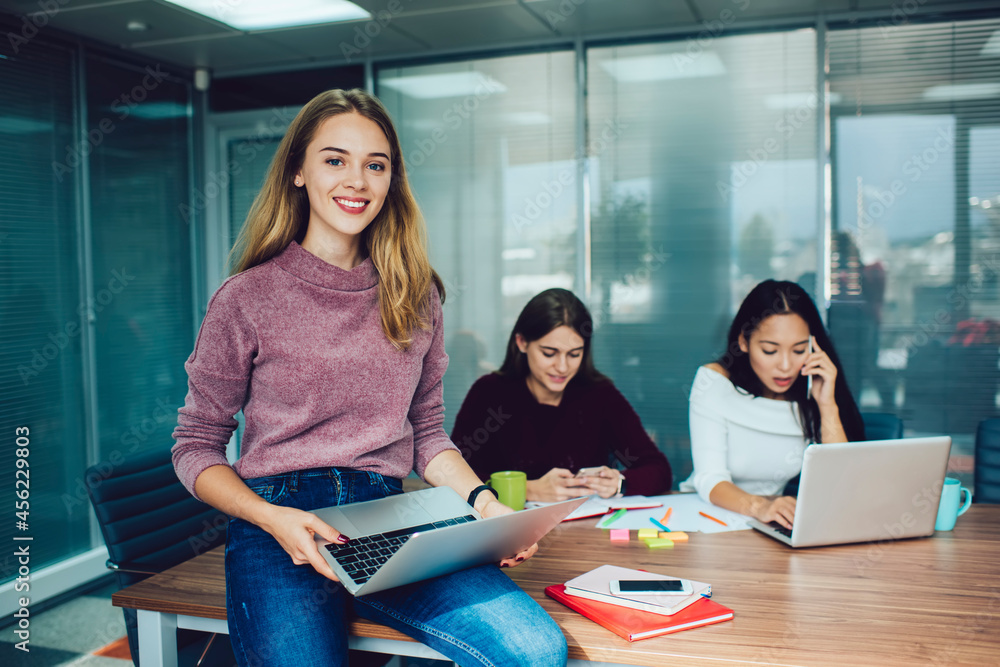 Poster portrait of cheerful female programmer holding digital laptop computer and smiling at camera during 
