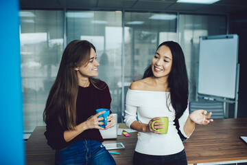 Cheerful Asian and Caucaisan colleagues enjoying gossip talking during work coffee break in office interior, happy diverse employees with caffeine beverage discussing workshop business and smiling - Powered by Adobe