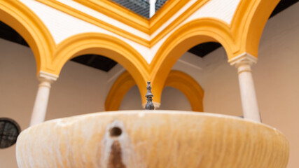 Iron faucet of a marble basin used as ornament in the middle of a patio, with yellow decorated arches. Seville Alcazar (meaning: fortress), Andalusia, Spain. Selective focus. Blurred background.