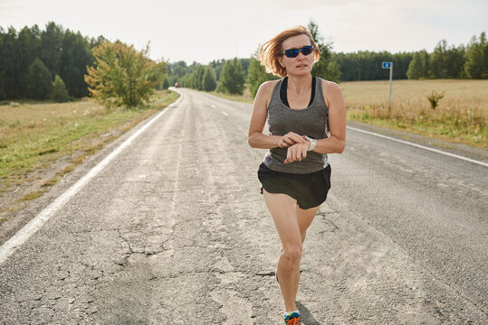 Mature Sportive Woman In Sports Clothing And In Sunglasses Running Along The Country Road