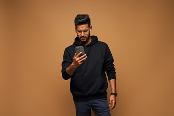 Handsome indian student in black hoody surfing his mobile phone on background