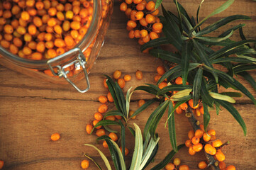 Sea buckthorn berries on a branch with green leaves on a wooden background. Glass jar with berries....