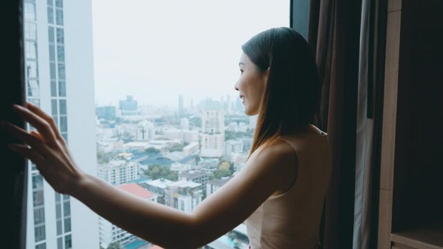 Asian woman open the curtain in hotel room to see the city view in luxury hotel