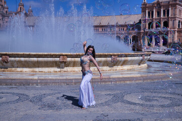 Obraz premium Middle-aged Hispanic woman, wearing a beautifully colored dress with rhinestones, for belly dancing, dancing surrounded by soap bubbles in front of a water fountain. Belly dance concept.