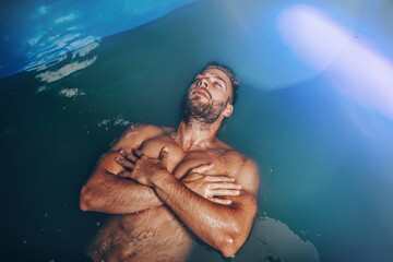 Handsome young bearded man floating in tank filled with dense salt water used in meditation, therapy, and alternative medicine.