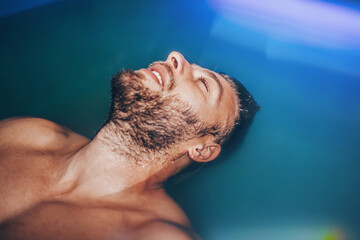 Handsome young bearded man floating in tank filled with dense salt water used in meditation, therapy, and alternative medicine.