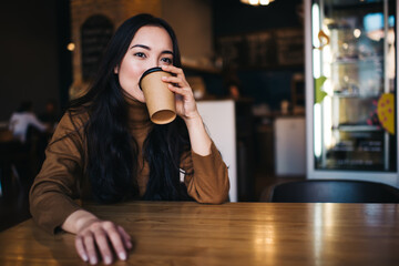 Pensive woman with brunette hair drinking hot caffeine beverage enjoying good taste in cafe interior, contemplative female thinking and dreaming during coffee break in cafeteria with wooden table