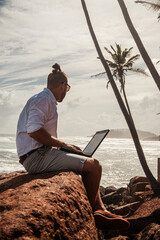 Fototapeta Cyfrowy nomada, człowiek pracujący z laptopem i smartfonem zdalnie na tle oceanu i skał siedzący na wybrzeżu. obraz